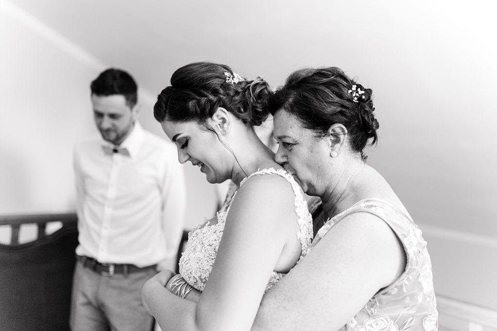 Pendant les préparatifs du mariage, la maman sert dans ses bras sa fille en tenue de mariée qui est de dos.